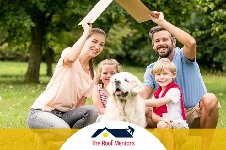 A cheerful family with two children and a dog sitting on the grass in a park, holding up a cardboard roof prop above their heads, with "the roof mentors" logo displayed below.