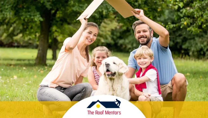 A cheerful family with two children and a dog sitting on the grass in a park, holding up a cardboard roof prop above their heads, with "the roof mentors" logo displayed below.