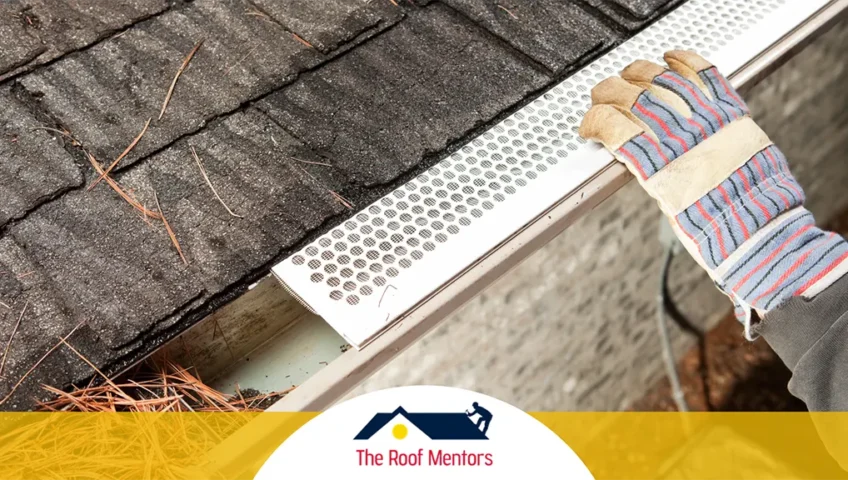 Worker's gloved hand installing gutter guards on a roof to prevent clogging by leaves, with the company logo of the roof mentors.